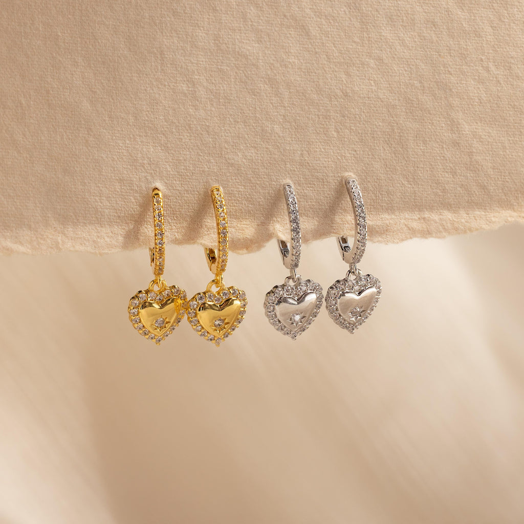 Pave Heart Drop Huggies in gold and silver, adorned with sparkling stones, displayed on a soft beige fabric background—an ideal romantic gift.