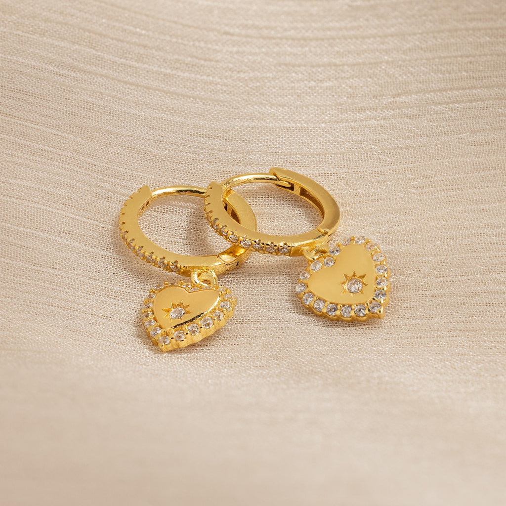 Pave Heart Drop Huggies, featuring heart-shaped pendants with sparkling clear stones, rest on beige textured fabric—an elegant and romantic gift.