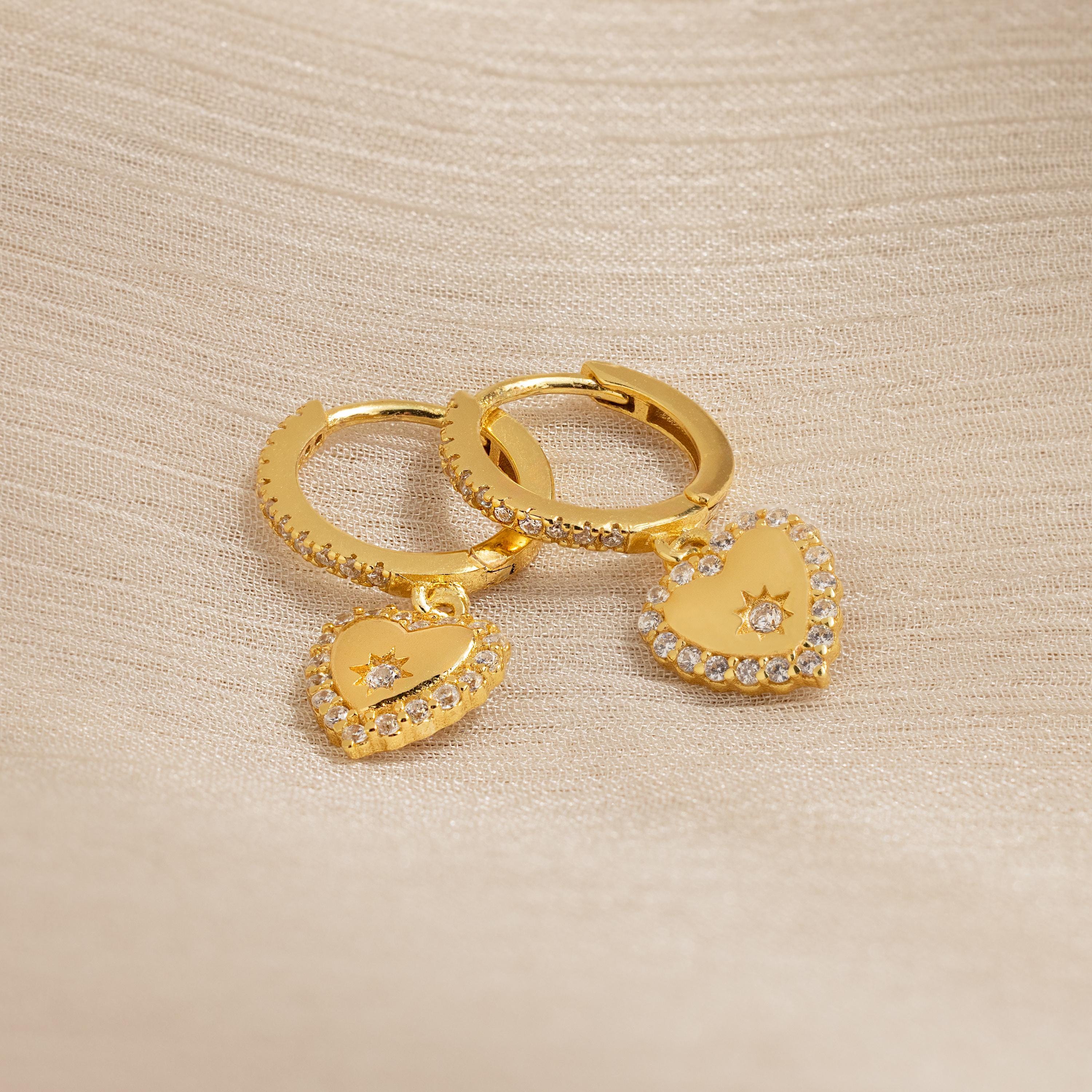 Pave Heart Drop Huggies, featuring heart-shaped pendants with sparkling clear stones, rest on beige textured fabric—an elegant and romantic gift.