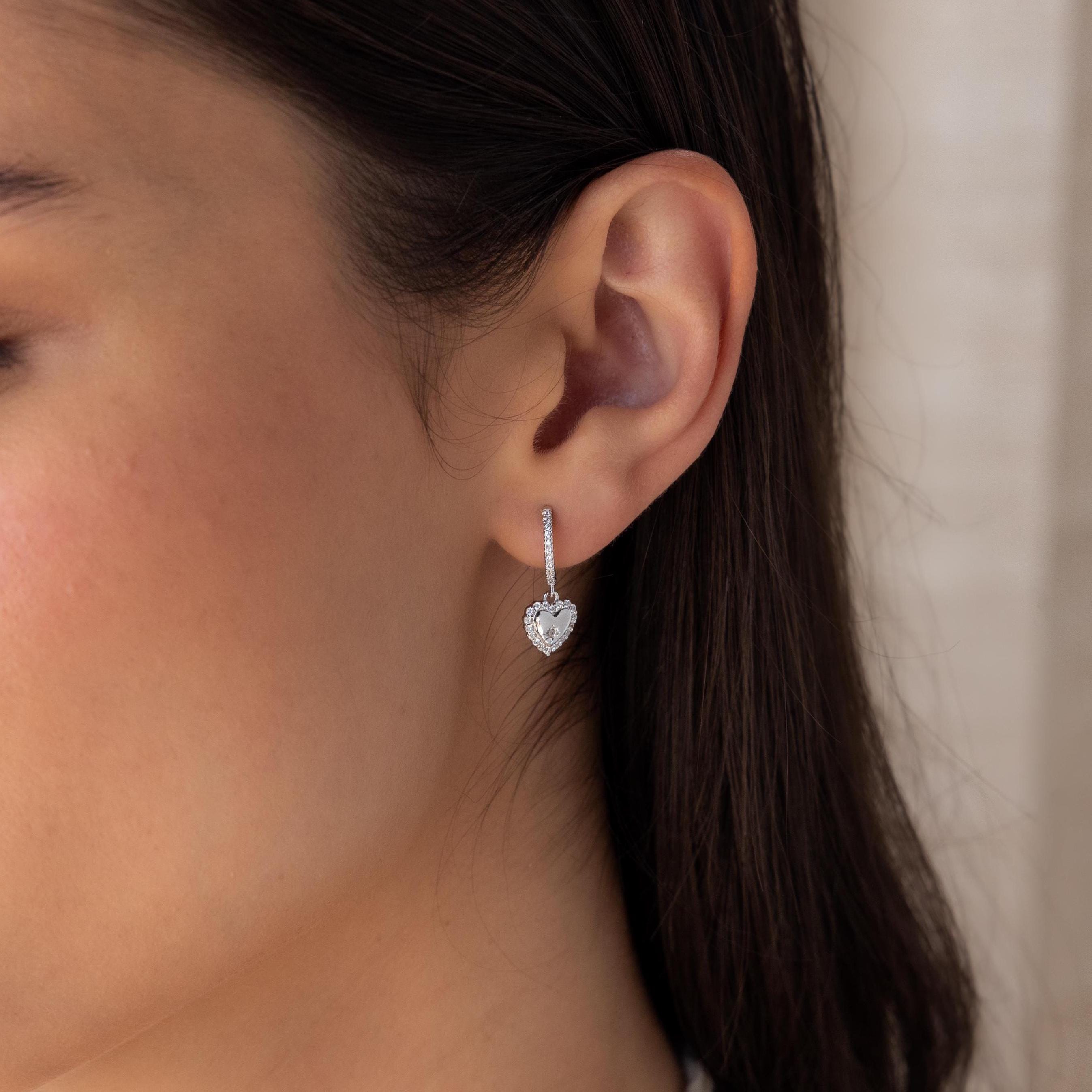 Close-up of a woman's ear wearing Pave Heart Drop Huggies—silver, heart-shaped huggie earrings adorned with sparkling gemstones, perfect as a romantic gift.