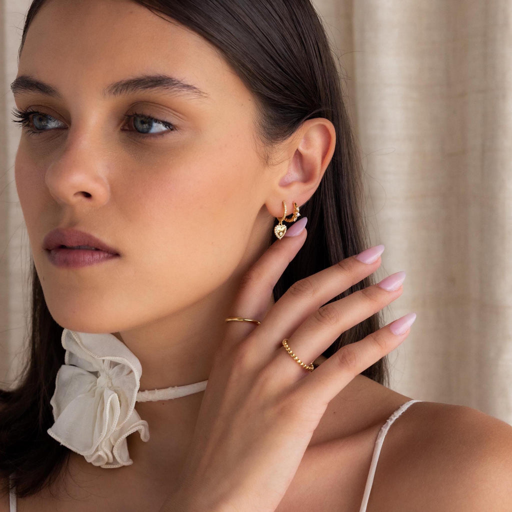 A woman wears gold jewelry, Pave Heart Drop Huggies, and a white ruffled choker, touching her ear with manicured fingers—an ideal romantic gift look.