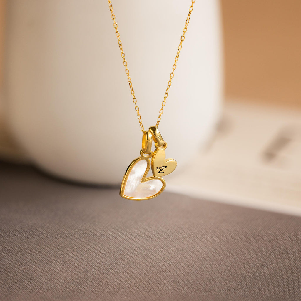 The Stylish Pearl Initial Heart Necklace features two pendants: a mother-of-pearl heart and an engraved letter "A," creating a personalized, meaningful accessory for any outfit.