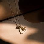 The Stylish Pearl Initial Heart Necklace in gold features open and small heart pendants, one engraved with an "R," displayed on a beige surface.