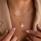 A woman wears layered gold necklaces, featuring the Stylish Pearl Initial Heart Necklace, over a beige blazer.