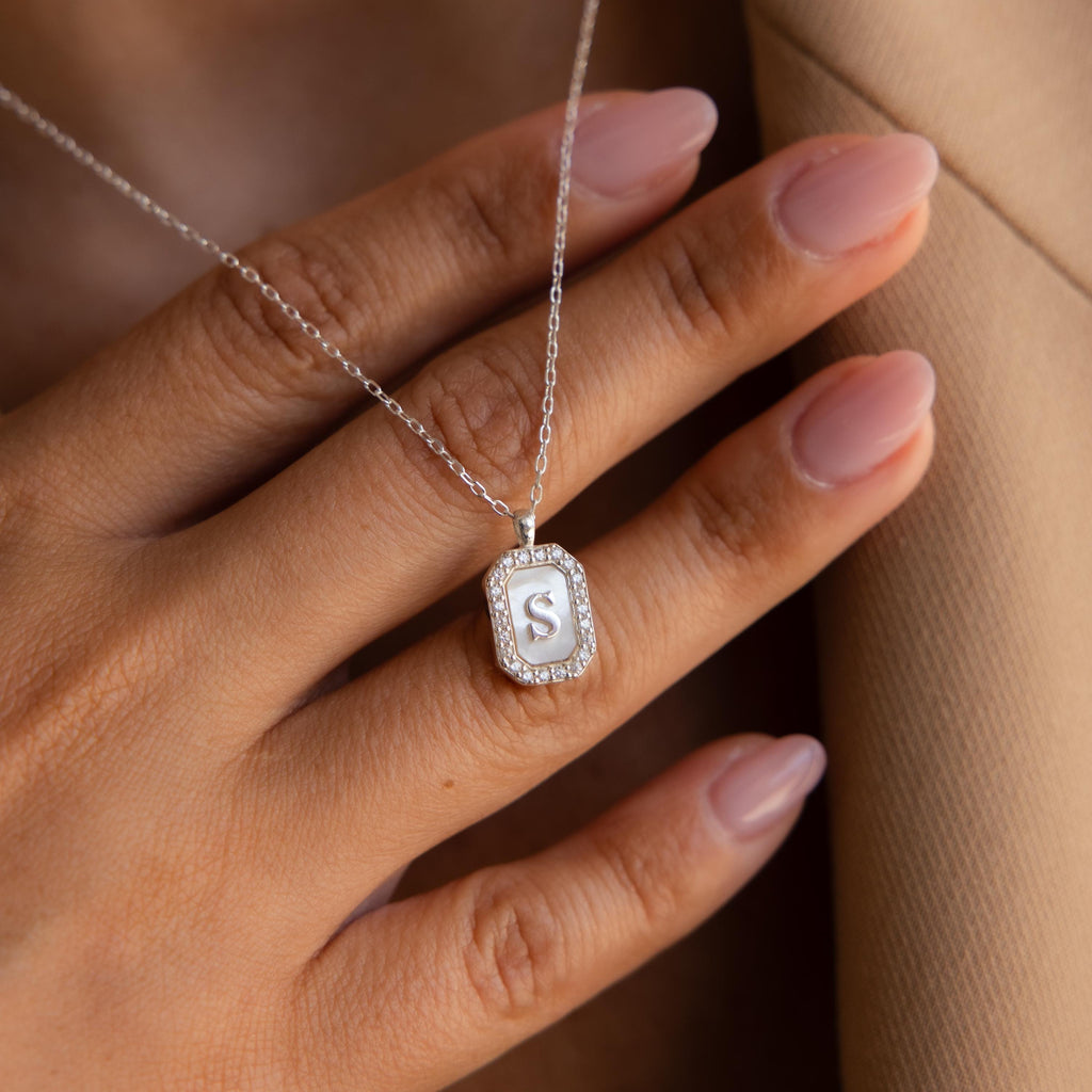 A hand with manicured nails holds the Pave Pearl Initial Necklace, its pendant featuring the letter S for a personalized touch.