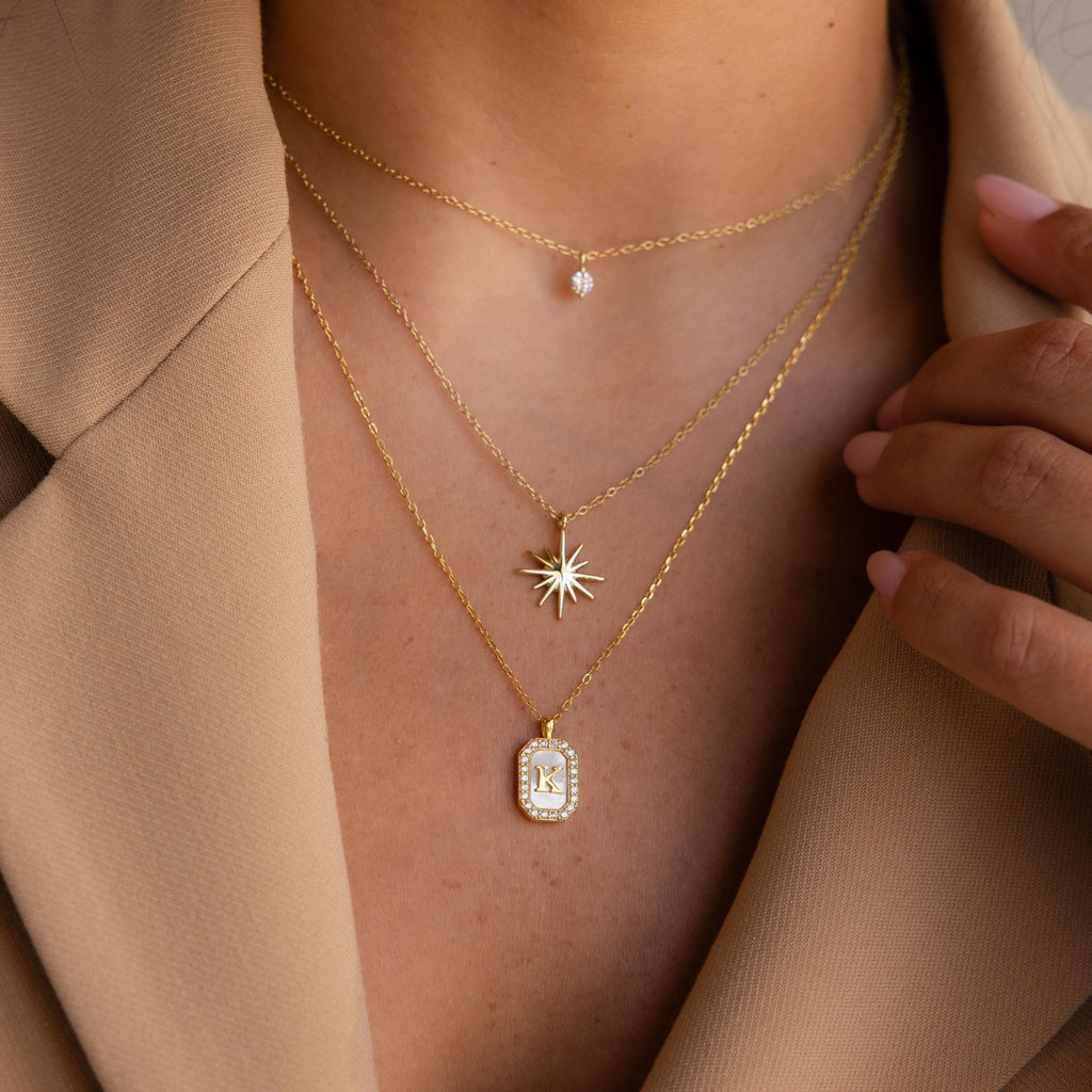 A woman in a beige blazer wears layered gold necklaces, featuring a CZ diamond necklace, pendants, and the Pave Pearl Initial Necklace.