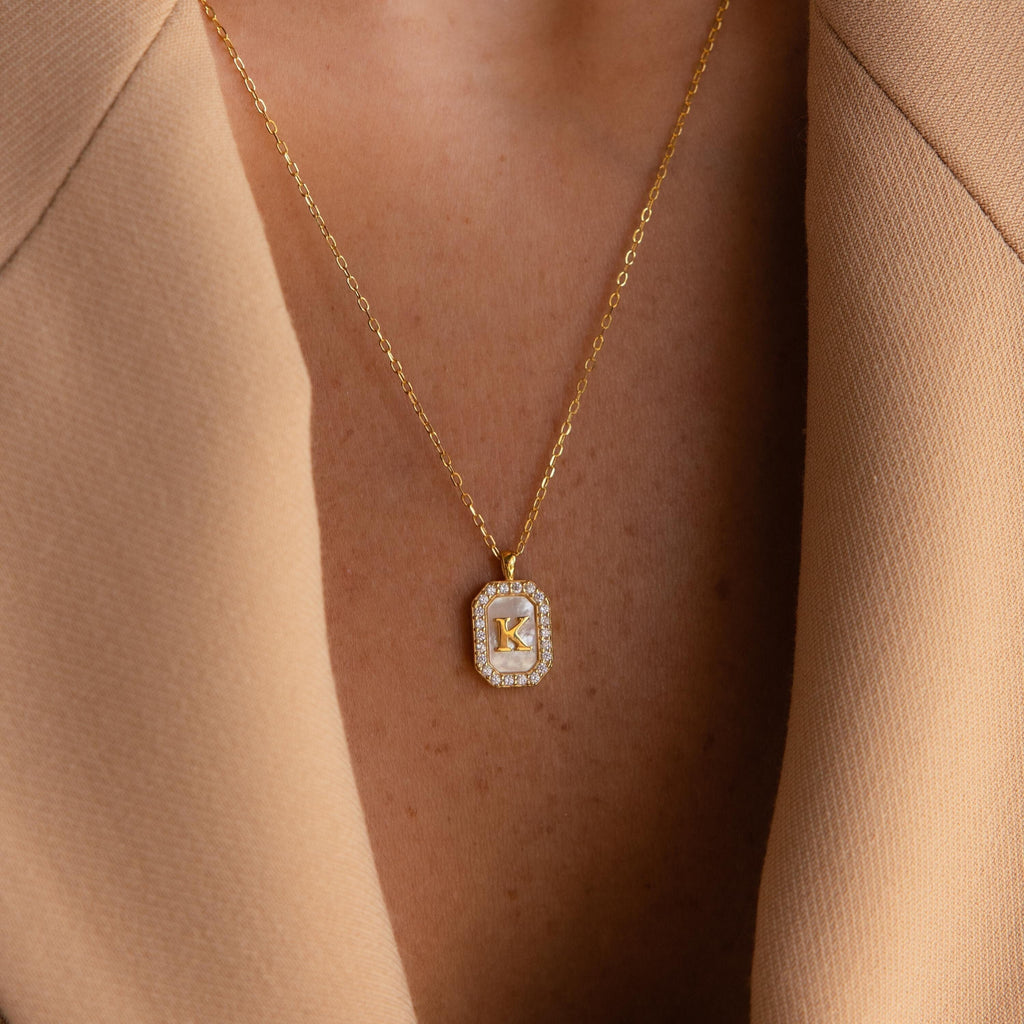 The Pave Pearl Initial Necklace, featuring a gold rectangular pendant with the letter K, is styled here with a beige blazer.