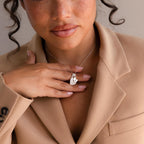 A woman in a beige blazer holds the Stylish Pearl Initial Heart Necklace to her chest, soft curls framing her face.