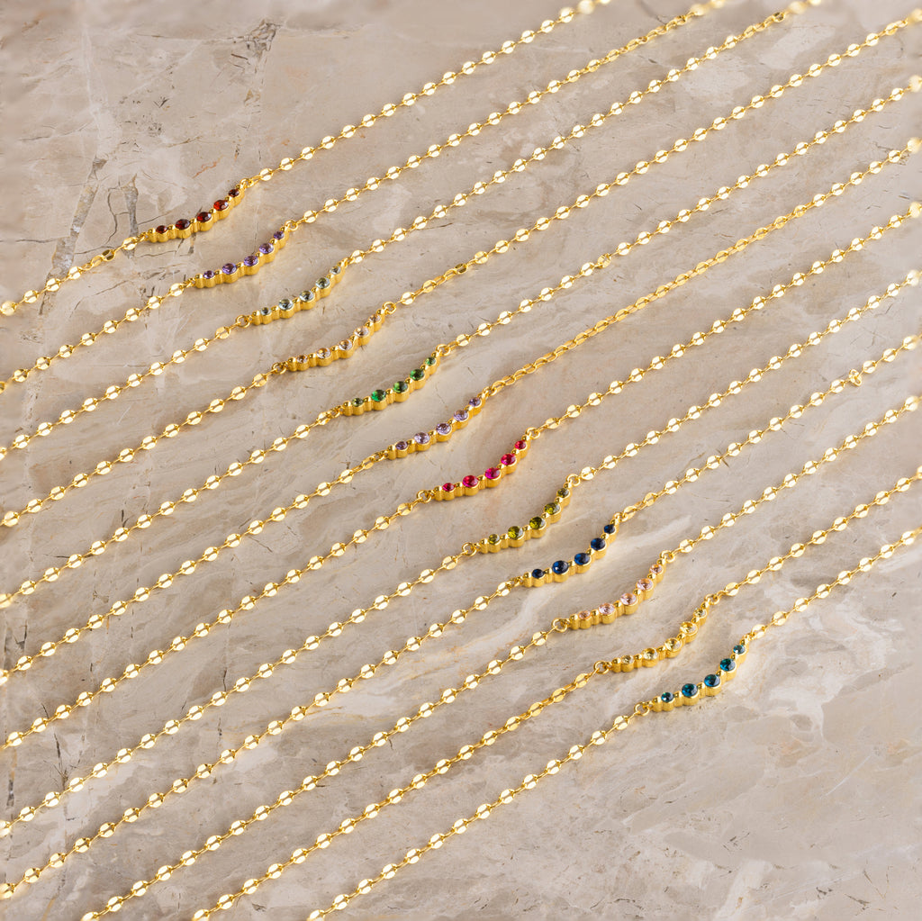 Nine Lace Birthstone Necklaces, each featuring a delicate gold chain and colorful gemstone, are arranged diagonally on a marble surface—perfect as dainty, personalized jewelry.