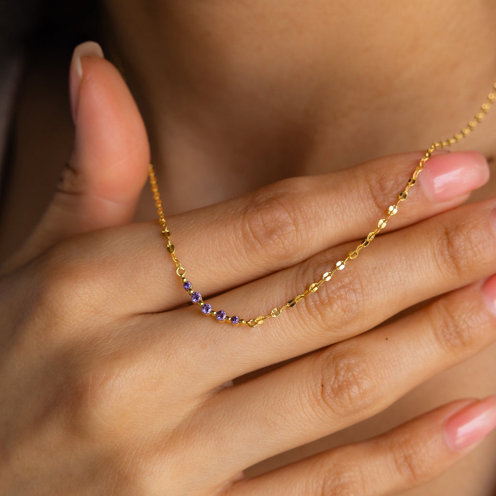 A hand with pink nails holds the Lace Birthstone Necklace, a dainty piece featuring small purple gemstones, close to the neck.