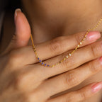 A hand with pink nails holds the Lace Birthstone Necklace, a dainty piece featuring small purple gemstones, close to the neck.