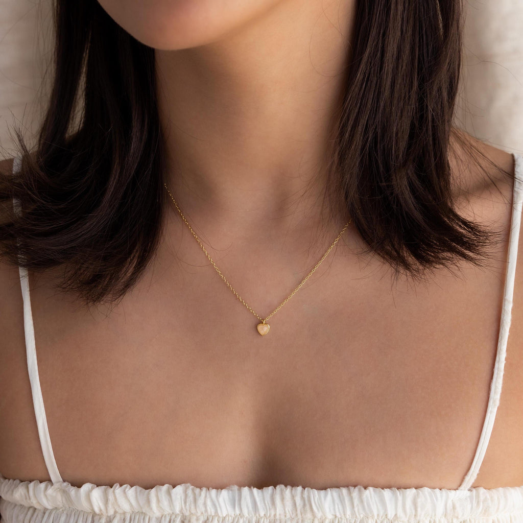 A woman wears the Tiny Rose Quartz Heart Necklace with a white top, pictured from shoulders to chin.
