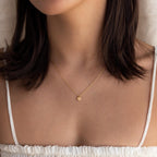 A woman wears the Tiny Rose Quartz Heart Necklace with a white top, pictured from shoulders to chin.