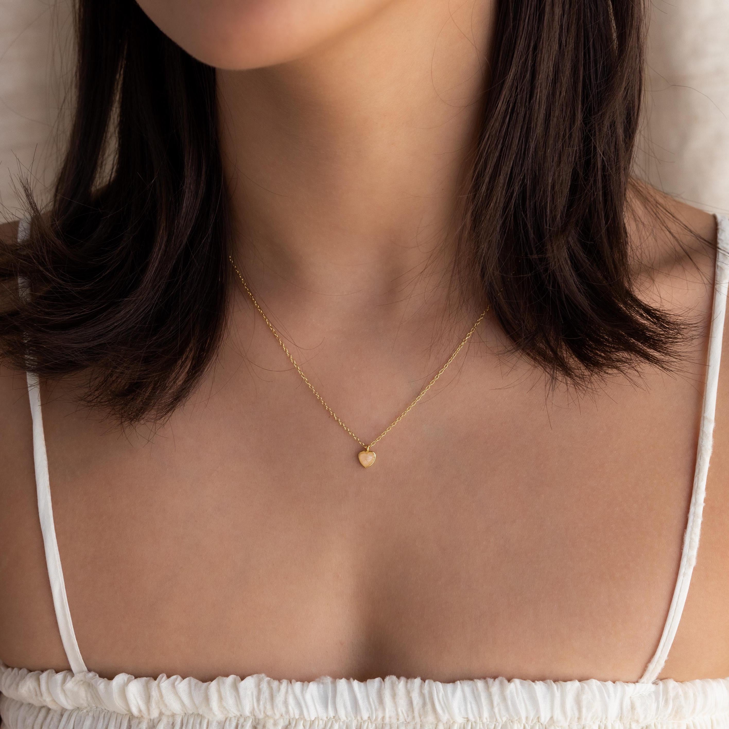 A woman wears the Tiny Rose Quartz Heart Necklace with a white top, pictured from shoulders to chin.