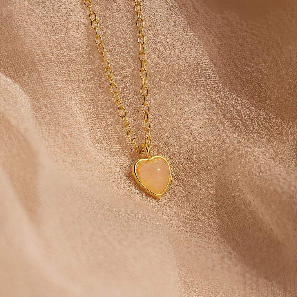 The Tiny Rose Quartz Heart Necklace features a delicate rose quartz heart pendant on a gold chain, beautifully displayed against a soft beige fabric background.