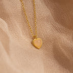 The Tiny Rose Quartz Heart Necklace features a delicate rose quartz heart pendant on a gold chain, beautifully displayed against a soft beige fabric background.