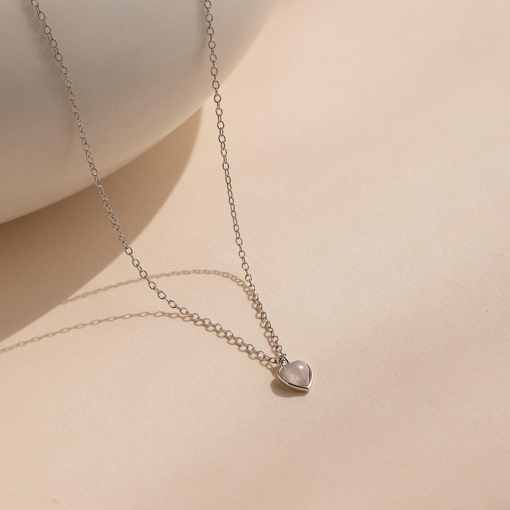 The Tiny Rose Quartz Heart Necklace features a delicate silver chain and a small rose quartz heart pendant, displayed on a light beige surface.