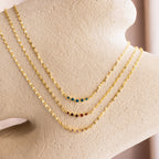 The Lace Birthstone Necklace features a dainty gold chain with a small colored gemstone, elegantly displayed on textured beige—perfect as a personalized or birthstone necklace for a unique, sophisticated touch.