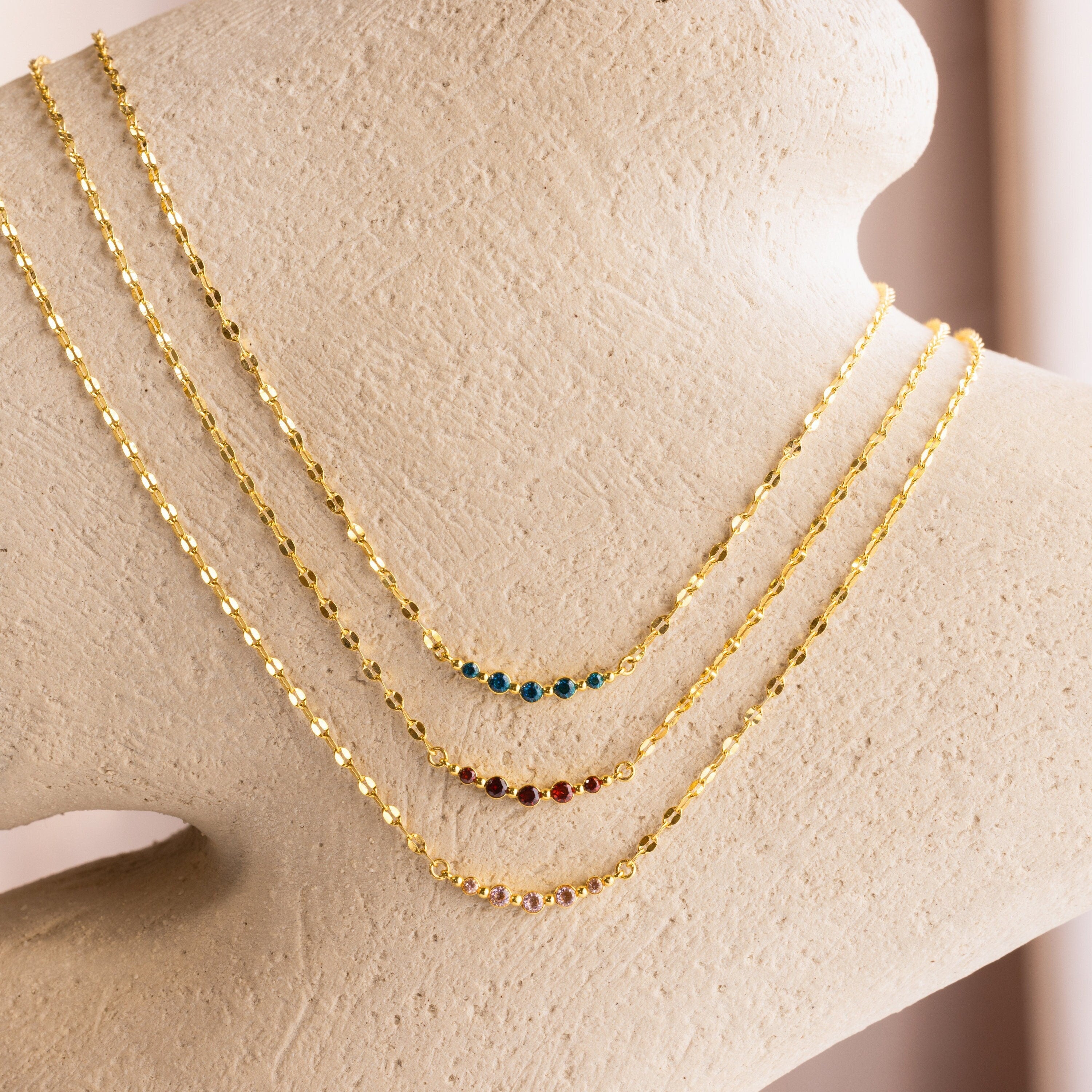 The Lace Birthstone Necklace features a dainty gold chain with a small colored gemstone, elegantly displayed on textured beige—perfect as a personalized or birthstone necklace for a unique, sophisticated touch.