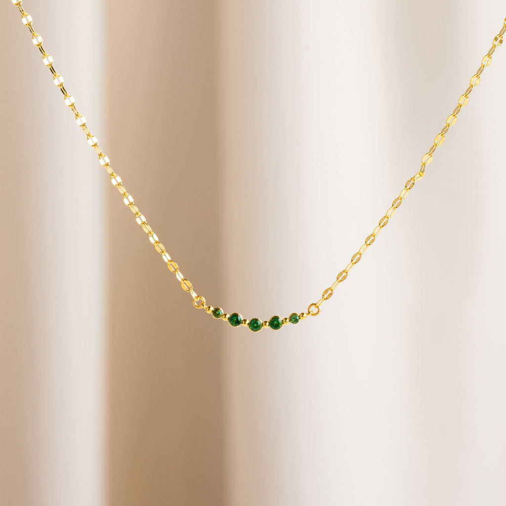 The Lace Birthstone Necklace features a gold chain with five small green beads, shown on a neutral light background—a delicate birthstone necklace ideal for everyday elegance.