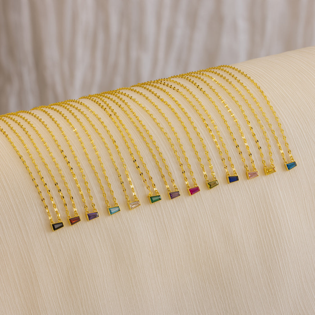 The Tapered Baguette Birthstone Necklace—a delicate gold mirror chain with a colorful rectangular gemstone pendant—is arranged neatly on a cream cloth.