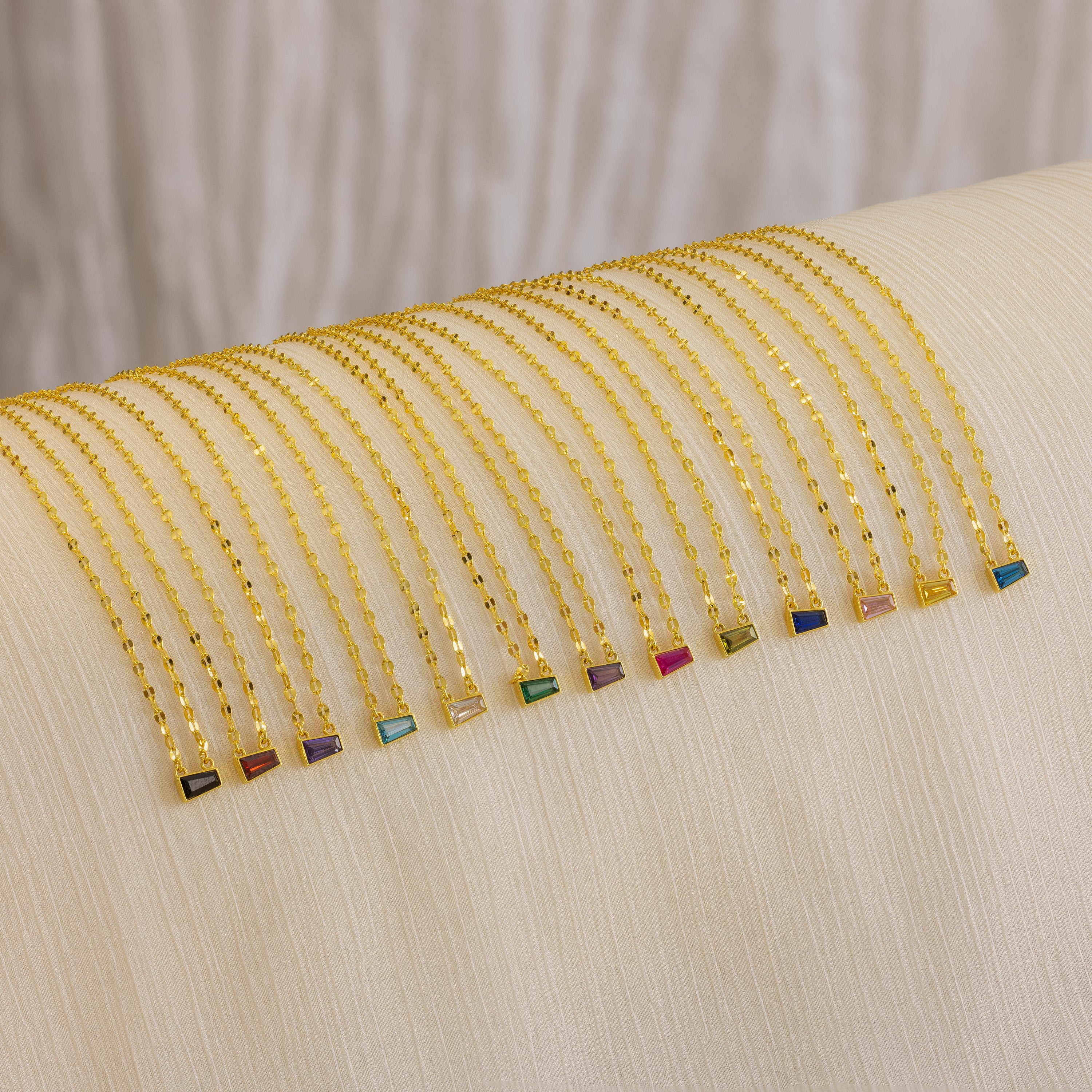 The Tapered Baguette Birthstone Necklace—a delicate gold mirror chain with a colorful rectangular gemstone pendant—is arranged neatly on a cream cloth.