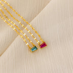 Two gold mirror chains with rectangular blue and pink pendants rest on cream fabric, showcasing the Tapered Baguette Birthstone Necklace for an elegant, personalized gemstone look.