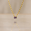 The Tapered Baguette Birthstone Necklace is a gold mirror chain necklace with a small rectangular purple gemstone pendant, photographed on a smooth surface.