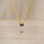 The Tapered Baguette Birthstone Necklace is a gold mirror chain necklace with a small rectangular purple gemstone pendant, photographed on a smooth surface.