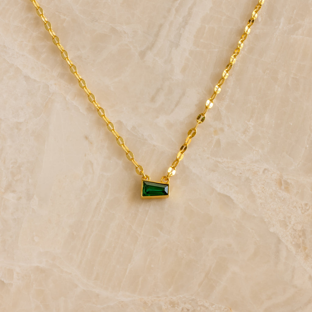 Tapered Baguette Birthstone Necklace: gold mirror chain with a small rectangular green pendant, shown on a beige marble background.
