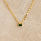 Tapered Baguette Birthstone Necklace: gold mirror chain with a small rectangular green pendant, shown on a beige marble background.