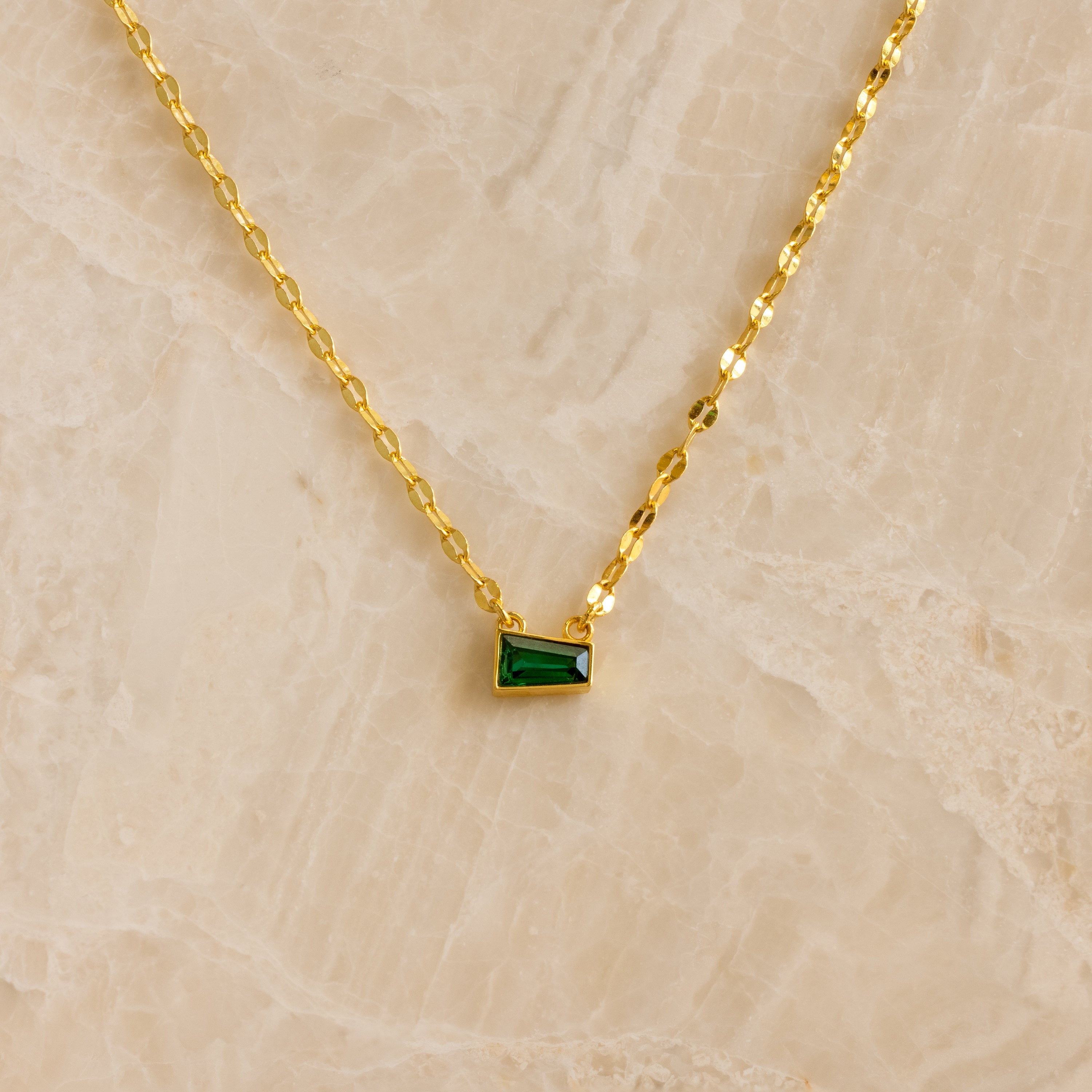 Tapered Baguette Birthstone Necklace: gold mirror chain with a small rectangular green pendant, shown on a beige marble background.