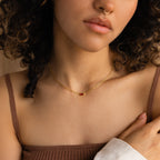 A woman with curly hair wears a Tapered Baguette Birthstone Necklace and a nose ring, dressed in a brown tank top.