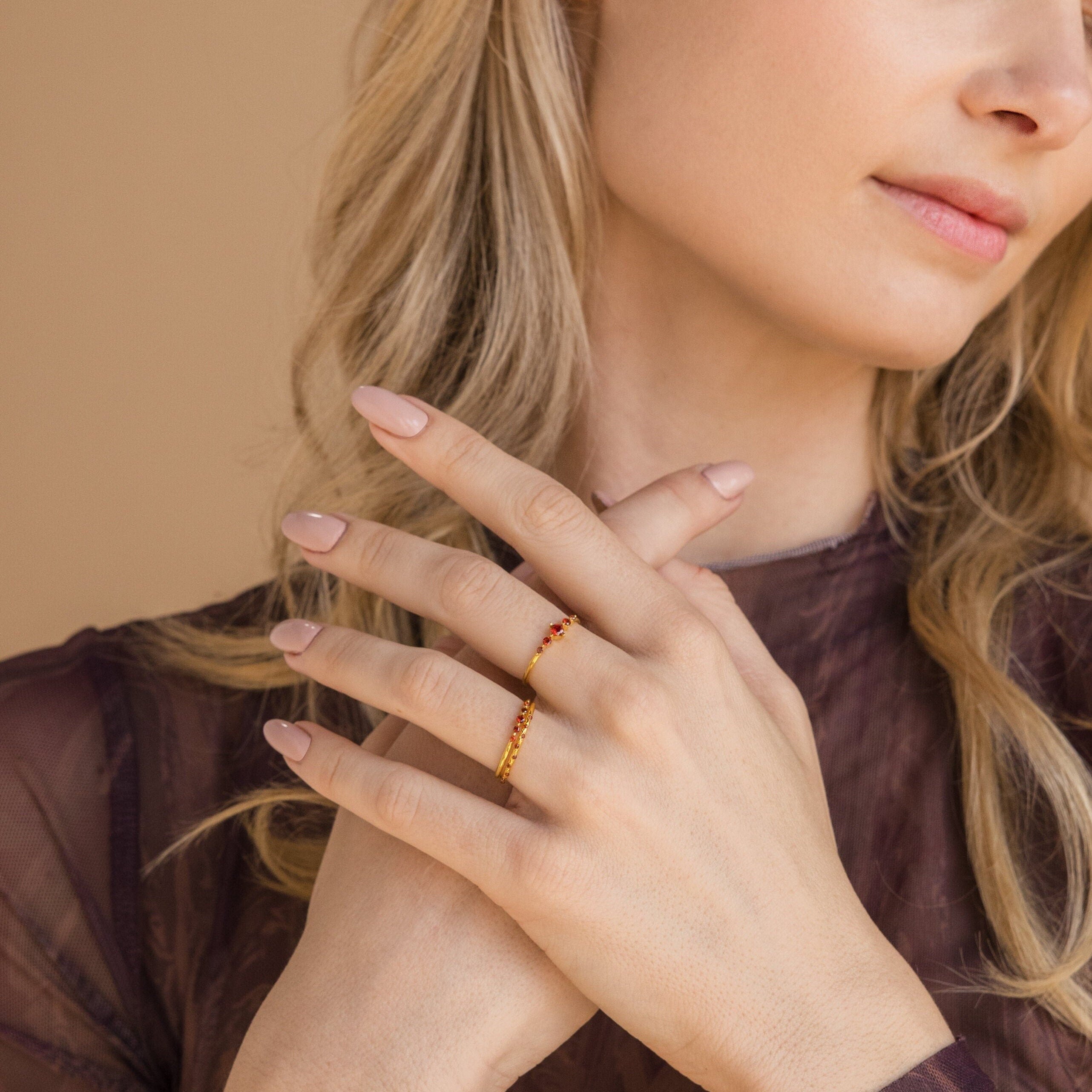 A woman wearing a sheer purple top showcases the Lace Birthstone Rings Set with small red stones on her finger—a thoughtful personalized gift.