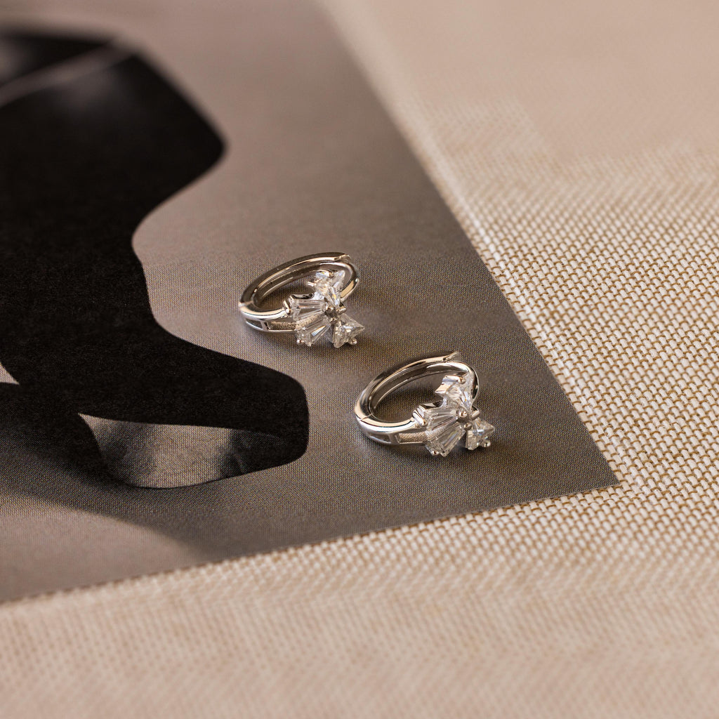 Close-up of Diamond Ribbon Bow Huggies in Silver set against a monochrome surface.