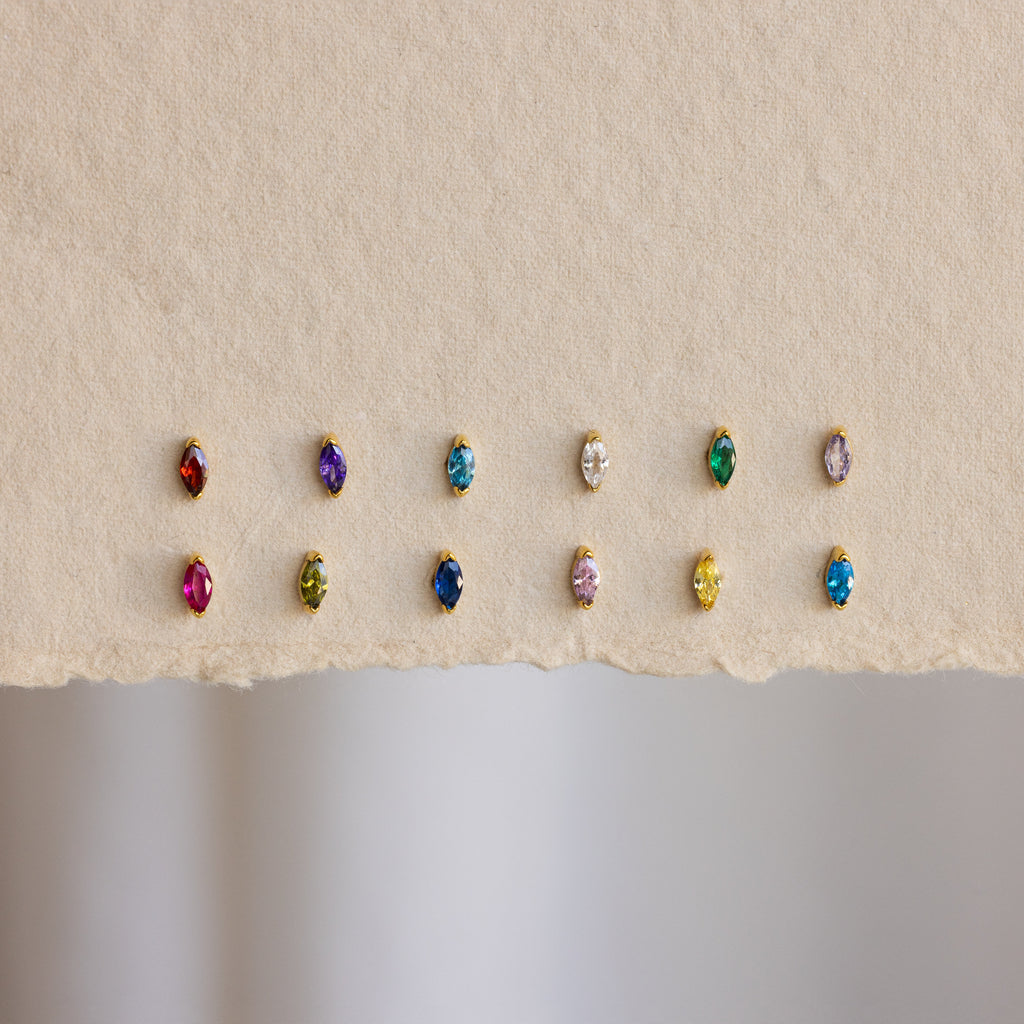 Twelve pairs of Marquise Birthstone Studs, each with colorful personalized gemstones in gold settings, are displayed on textured paper.