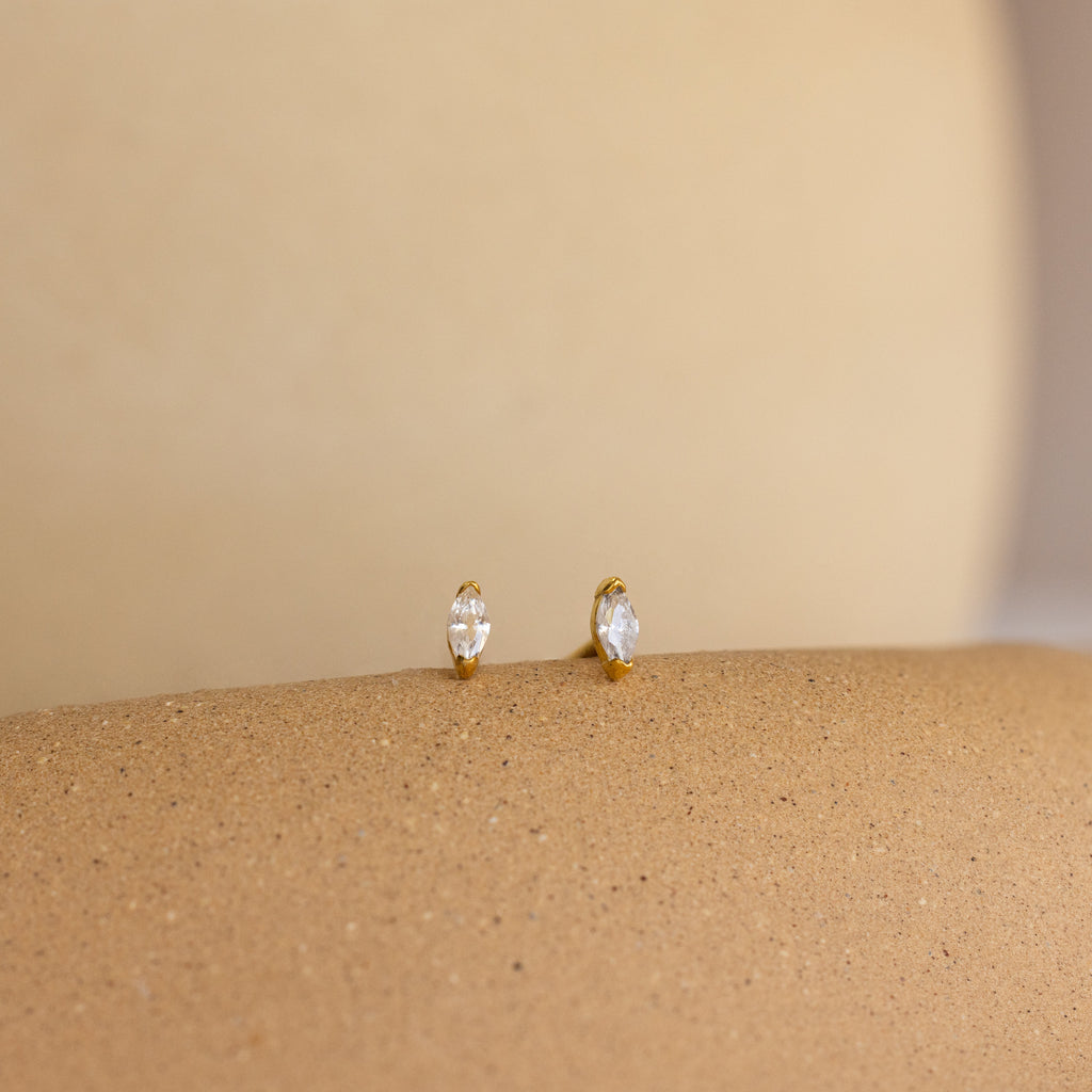 Two Marquise Birthstone Studs with clear gems rest on a smooth, beige textured surface.