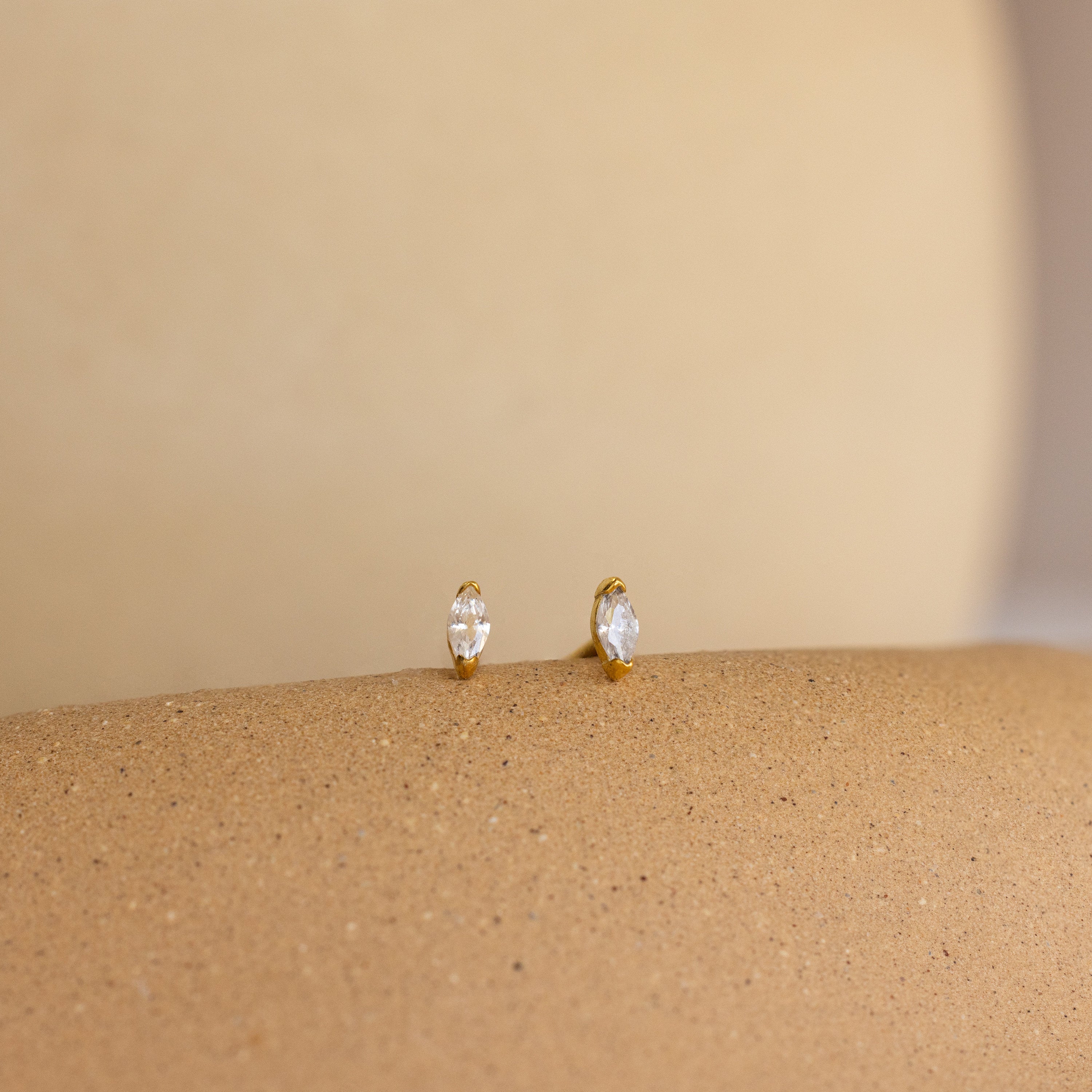 Two Marquise Birthstone Studs with clear gems rest on a smooth, beige textured surface.