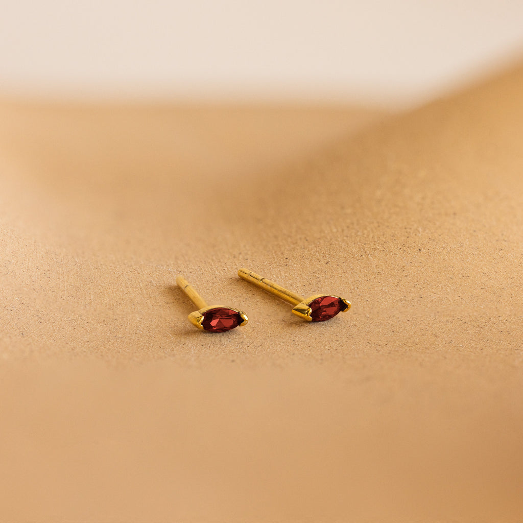 Two gold marquise-cut stud earrings with vibrant red gemstones from our Marquise Birthstone Studs collection, perfect for adding a personalized touch to your look. Displayed on a sandy beige surface.