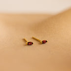 Two gold marquise-cut stud earrings with vibrant red gemstones from our Marquise Birthstone Studs collection, perfect for adding a personalized touch to your look. Displayed on a sandy beige surface.