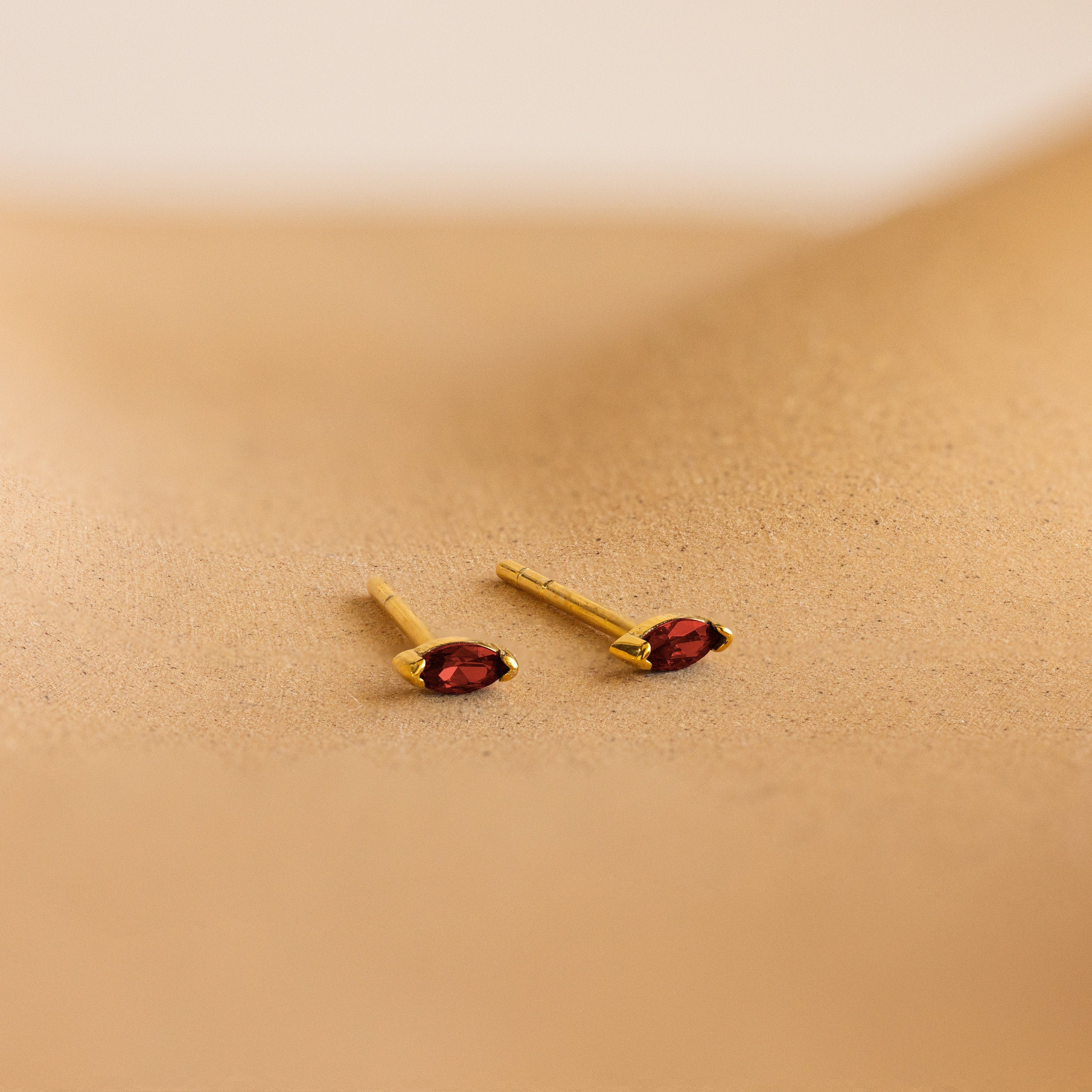 Two gold marquise-cut stud earrings with vibrant red gemstones from our Marquise Birthstone Studs collection, perfect for adding a personalized touch to your look. Displayed on a sandy beige surface.
