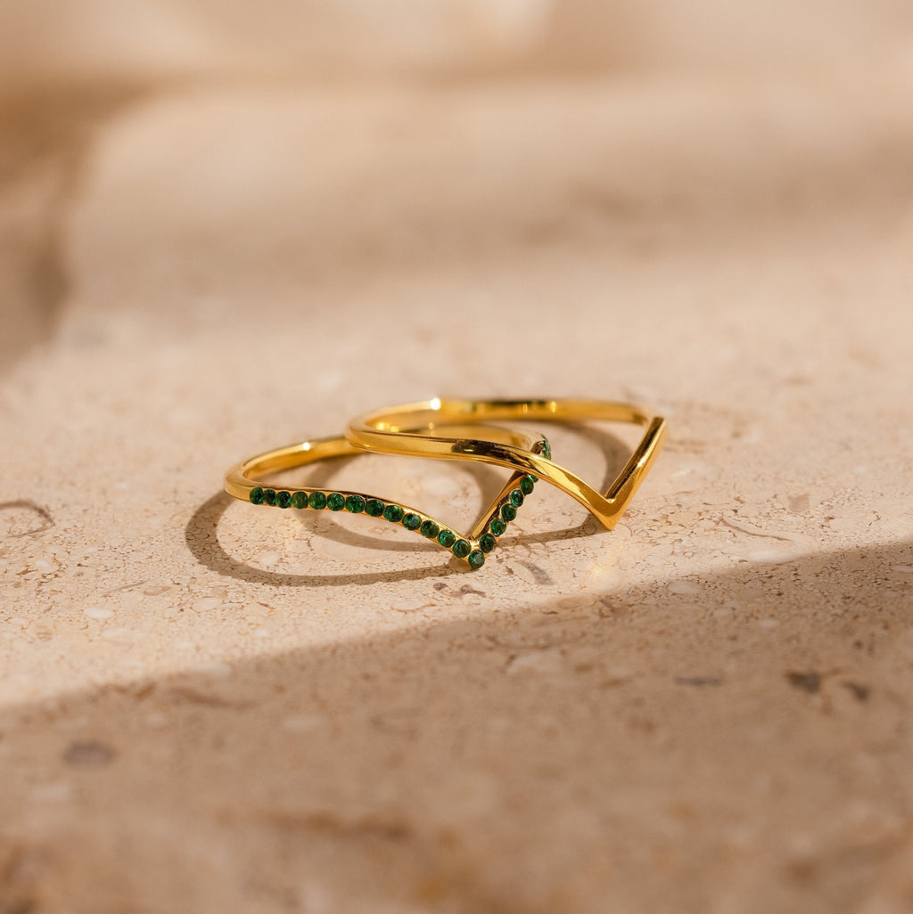 Two Chevron Birthstone Rings Set with green gemstones are displayed on a beige stone surface in soft, natural light.