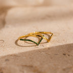 Two Chevron Birthstone Rings Set with green gemstones are displayed on a beige stone surface in soft, natural light.