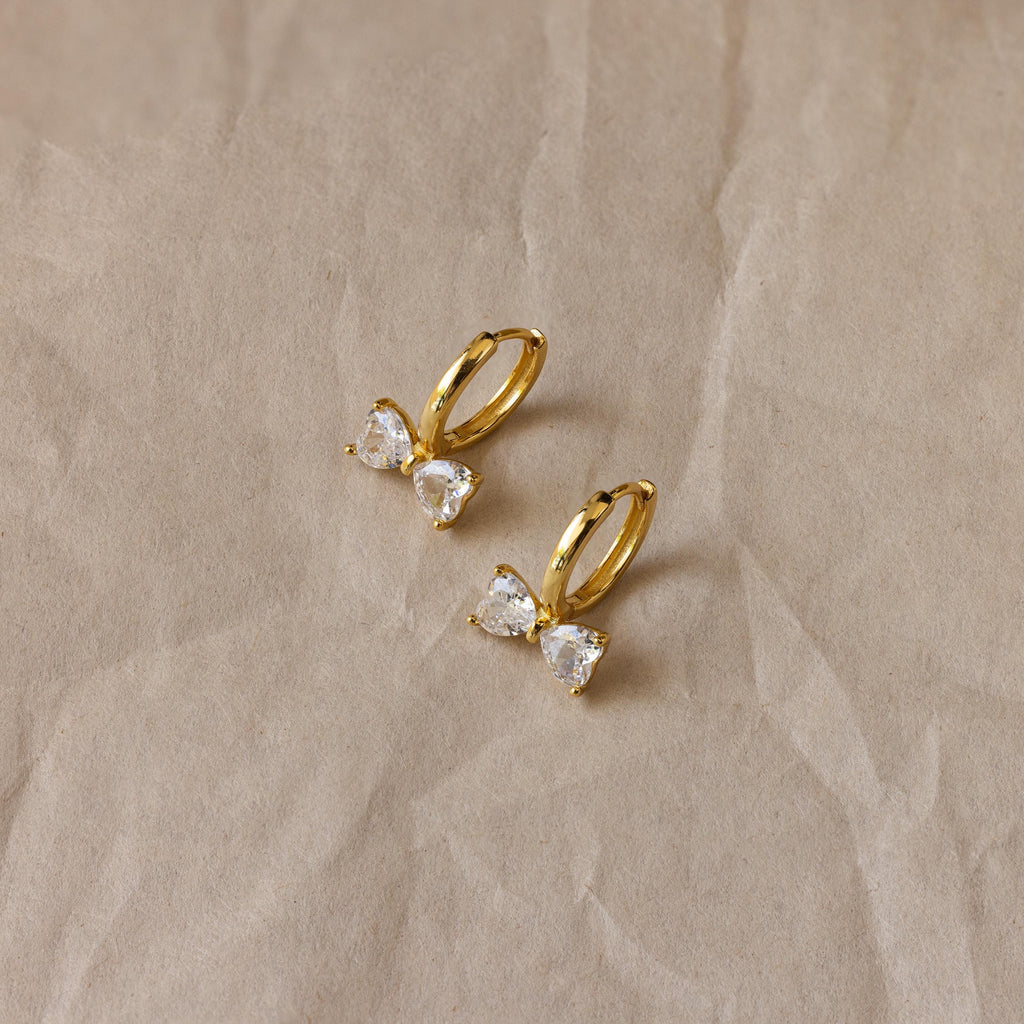 Diamond Bow Huggies, gold hoop earrings inspired by delicate bowties and adorned with sparkling gemstones, displayed on a crumpled beige background.