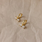 Diamond Bow Huggies, gold hoop earrings inspired by delicate bowties and adorned with sparkling gemstones, displayed on a crumpled beige background.