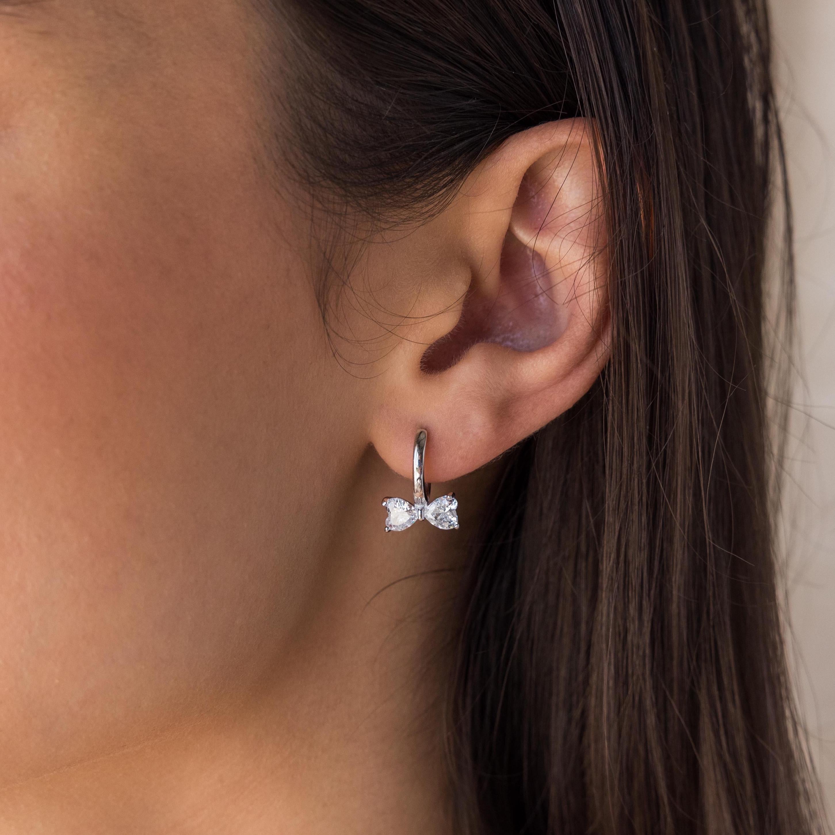 Close-up of a woman's ear wearing Diamond Bow Huggies—silver hoop earrings featuring a delicate bow-shaped design.