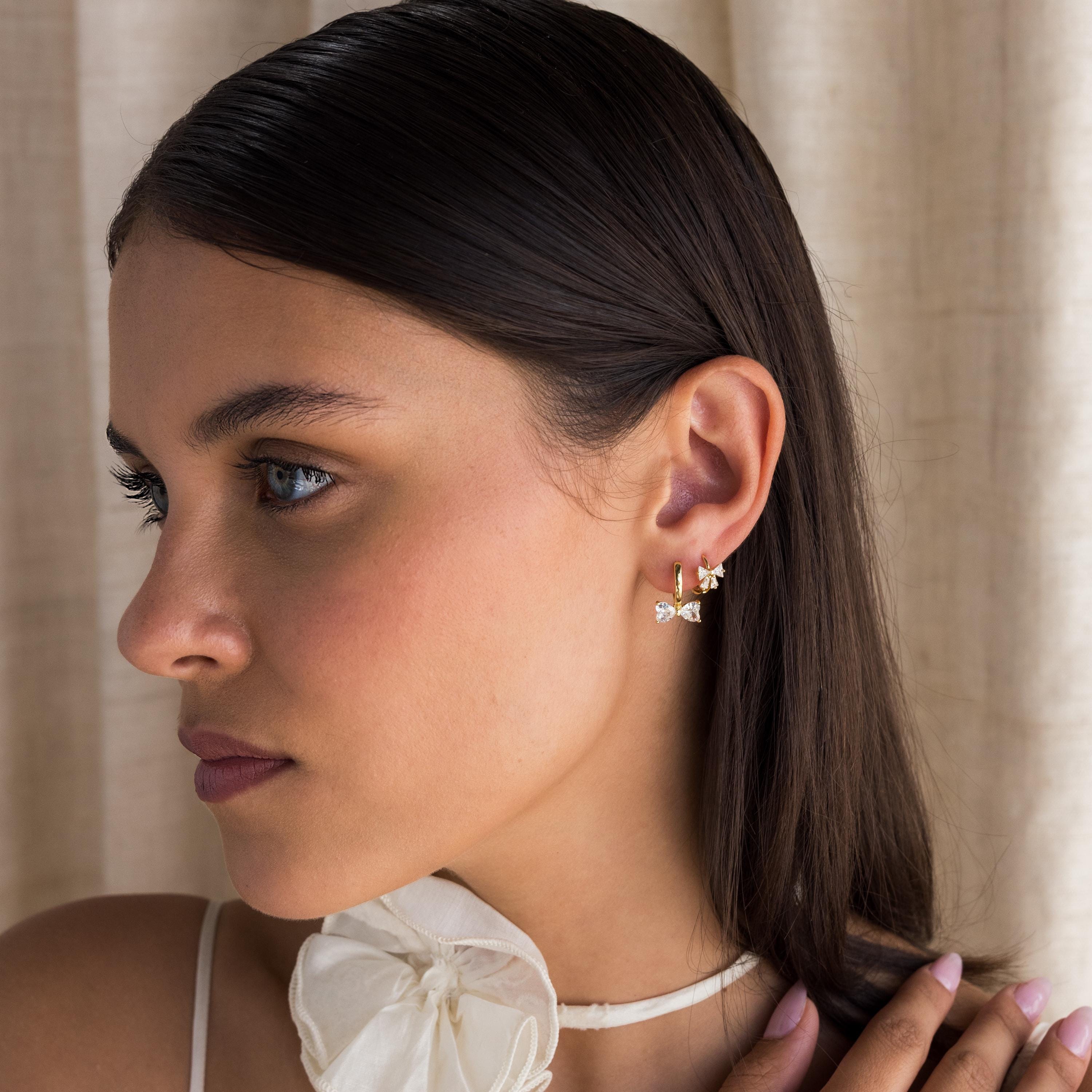 A woman with long dark hair looks to the left, wearing Diamond Bow Huggies and a white bow choker.
