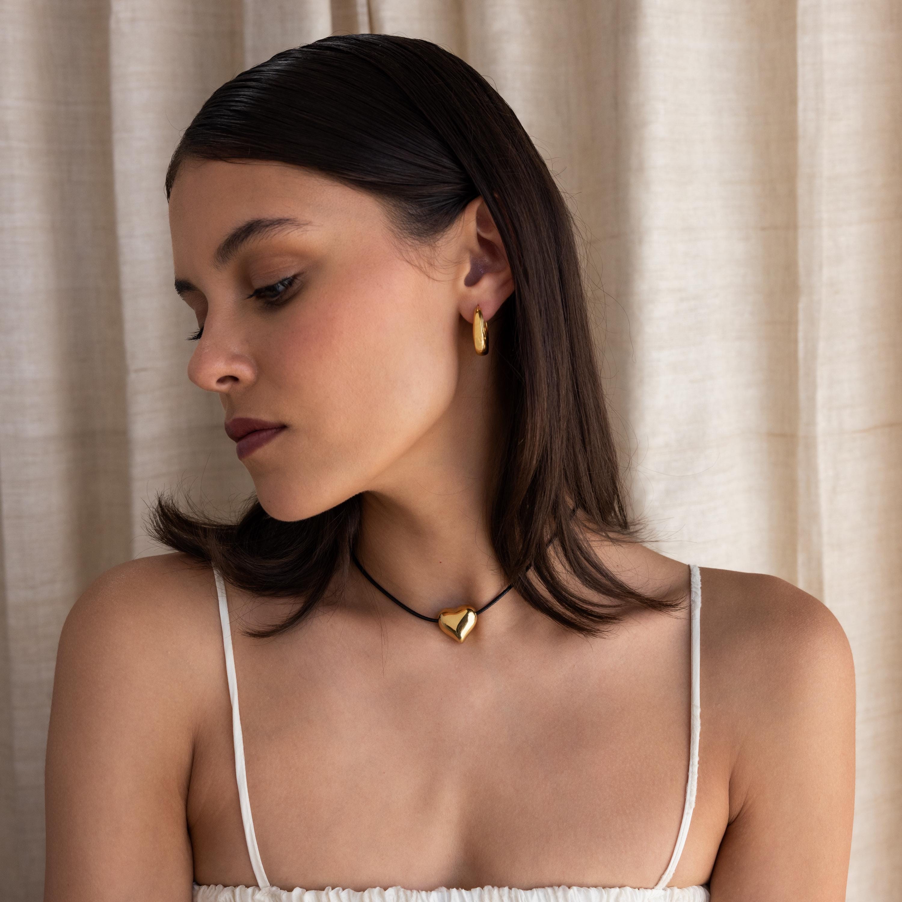 A woman with dark hair wears gold earrings and the Chunky Puff Heart Necklace, looking down.