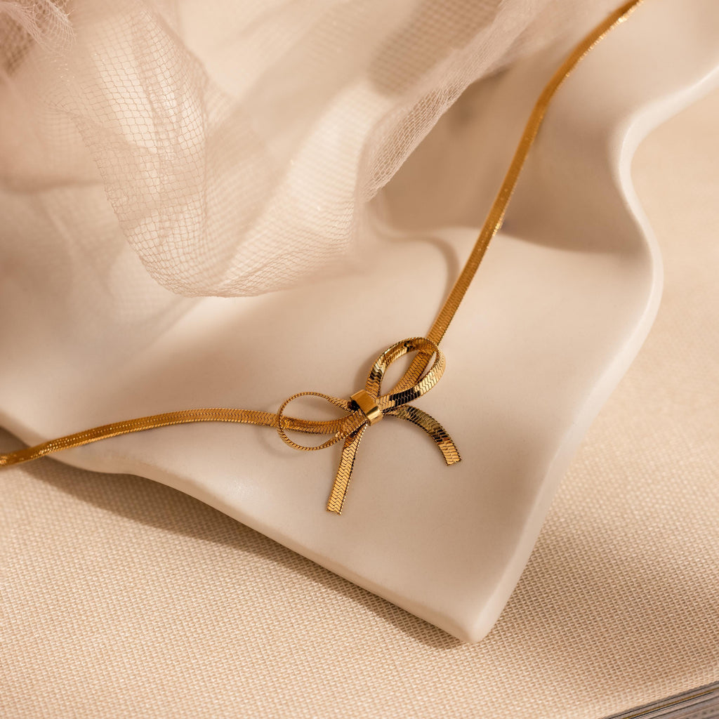 The Herringbone Bow Necklace in 18K Gold, styled in a coquette fashion, is showcased on a cream wavy ceramic dish with soft fabric in the background.