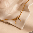 The Herringbone Bow Necklace in 18K Gold, styled in a coquette fashion, is showcased on a cream wavy ceramic dish with soft fabric in the background.